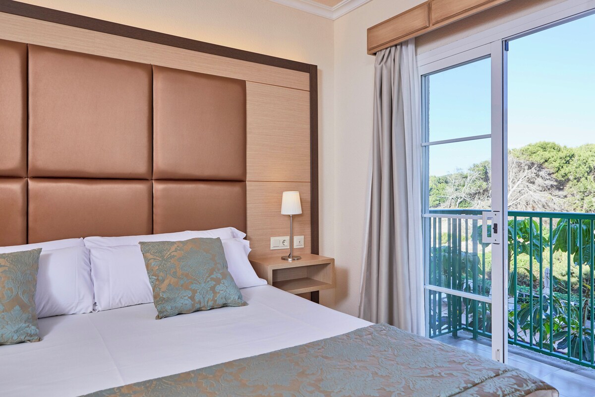 A hotel room with a large bed, a nightstand with a lamp, and a view of a balcony with greenery.