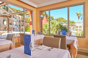 A restaurant with tables set for dining, a menu stand, and large windows offering a view of buildings and trees. - Zafiro Cala Mesquida (Capdepera)