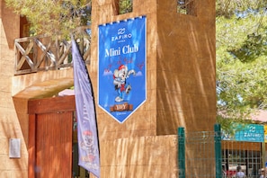A Zafiro Hotels Mini Club sign with a cartoon character, a wooden door, and a green fence.