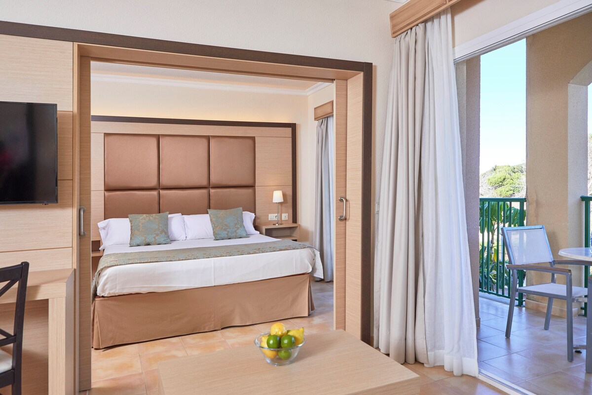 A hotel room with a bed, a TV, a table with fruit, and a balcony with chairs.