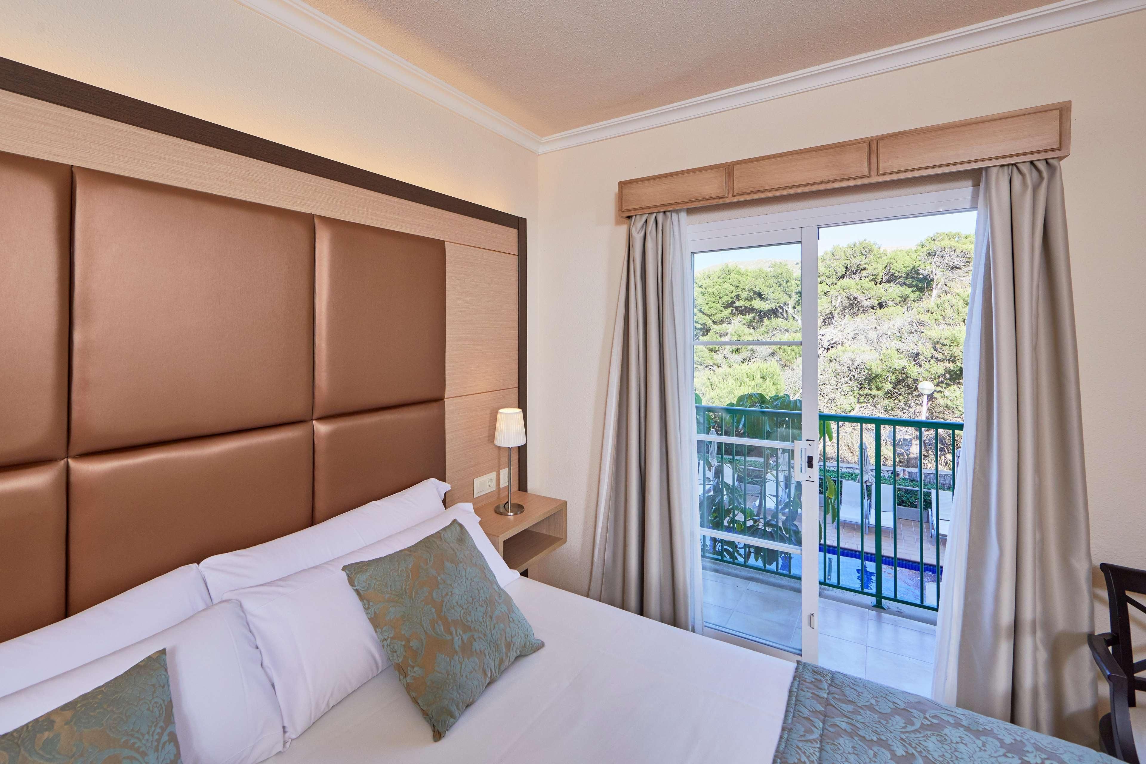 A hotel room with a large bed, a nightstand, a lamp, and a view of a balcony with greenery.