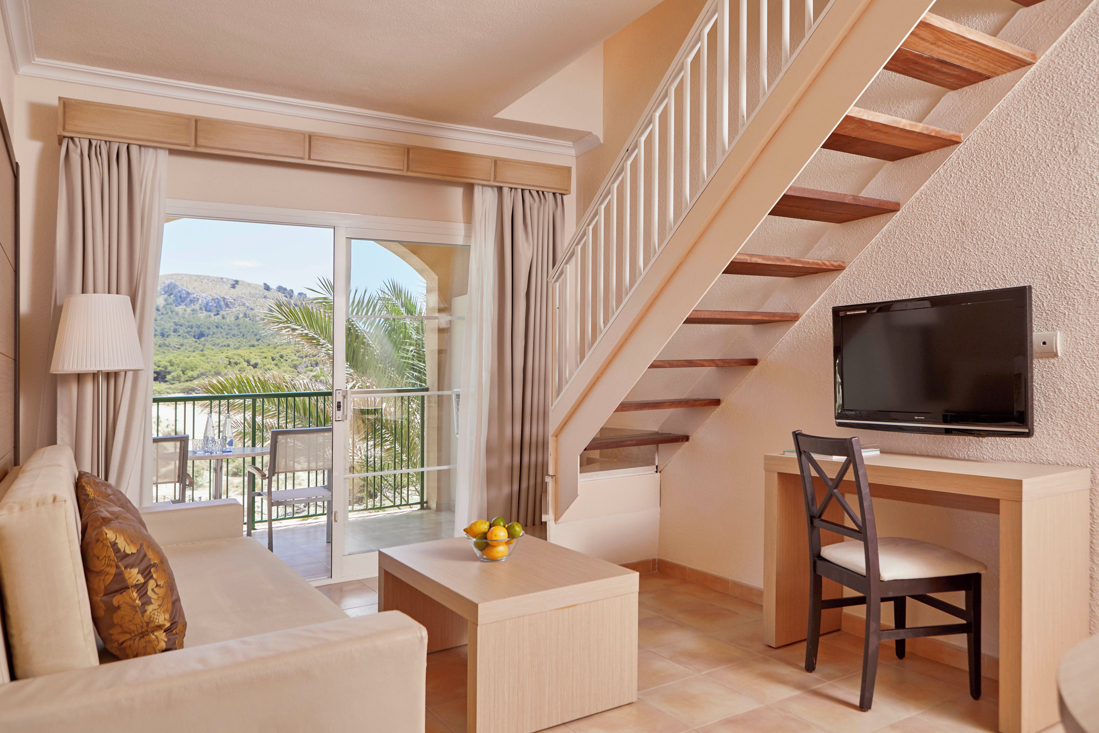 A living room with a sofa, a coffee table, a TV, and a desk with a chair. There is a balcony with a view of greenery and a mountain.
