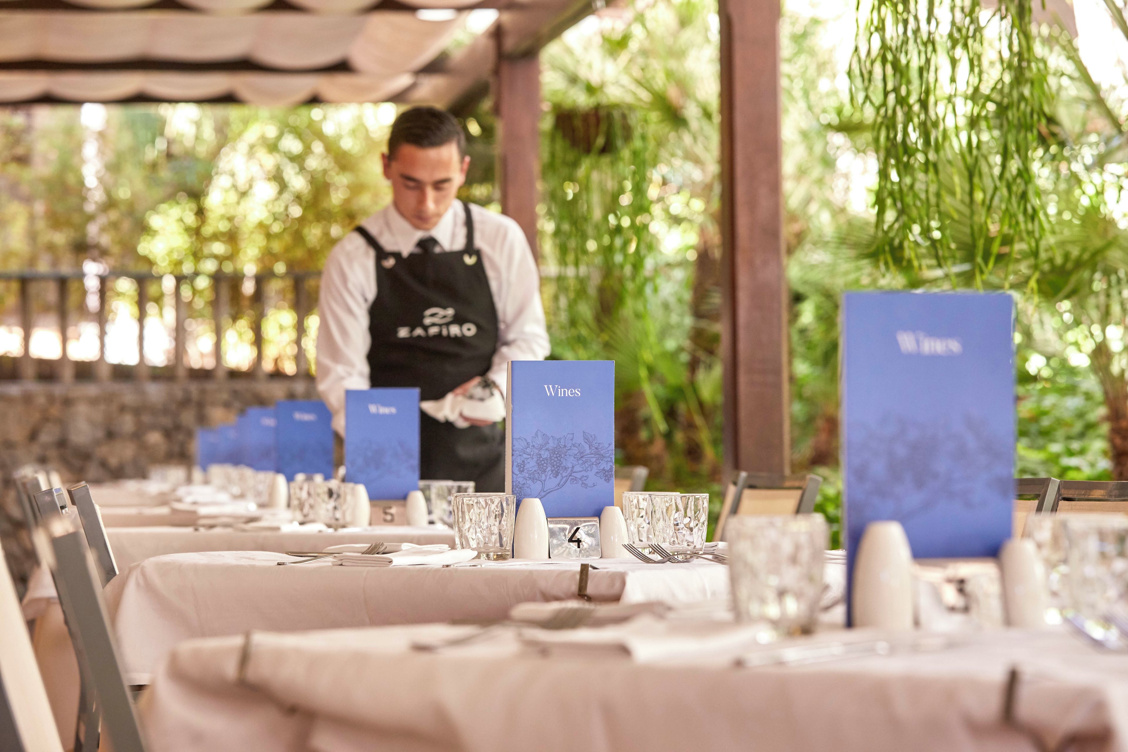 A restaurant setting with tables set for dining, a waiter in uniform, and wine menu cards.