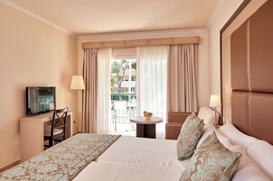 Habitación de hotel con cama, escritorio con televisor, pequeña mesa redonda con frutero y balcón con silla y mesa.