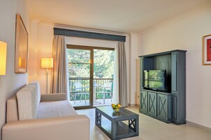 Una sala de estar moderna con un sofá, una mesa de centro con fruta, un televisor sobre un mueble bajo y un balcón con sillas y cortinas.