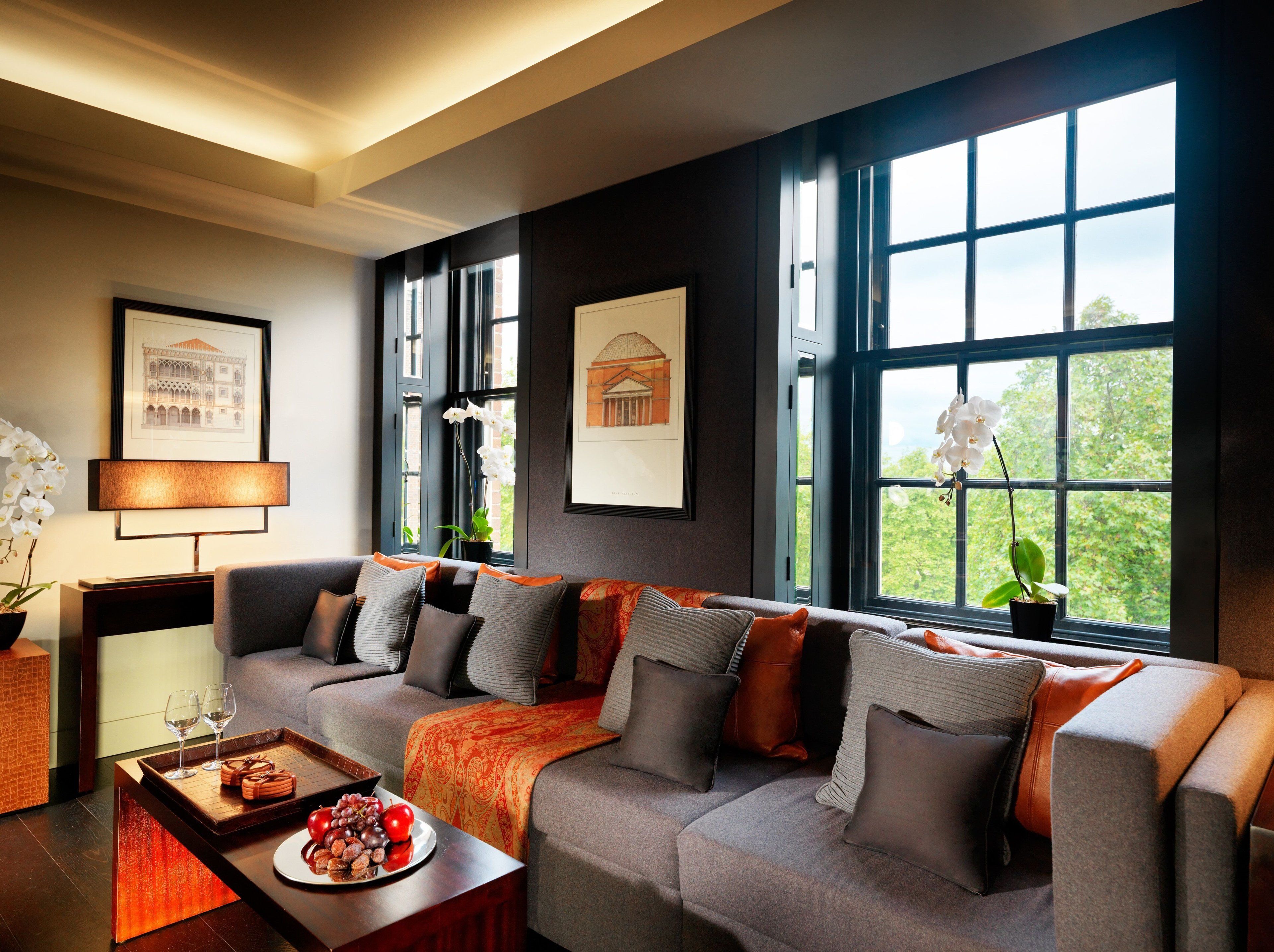 Un salon moderne avec un canapé gris, un plaid orange et une table à manger sur laquelle est disposée une assiette de fruits.