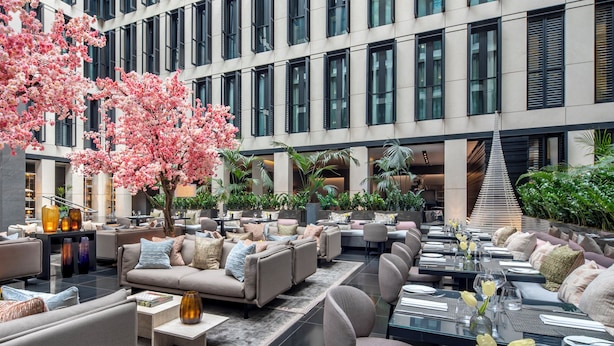 A modern indoor dining area with multiple seating arrangements, including sofas and tables, and a large tree with pink blossoms. - Grosvenor House Suites (London)