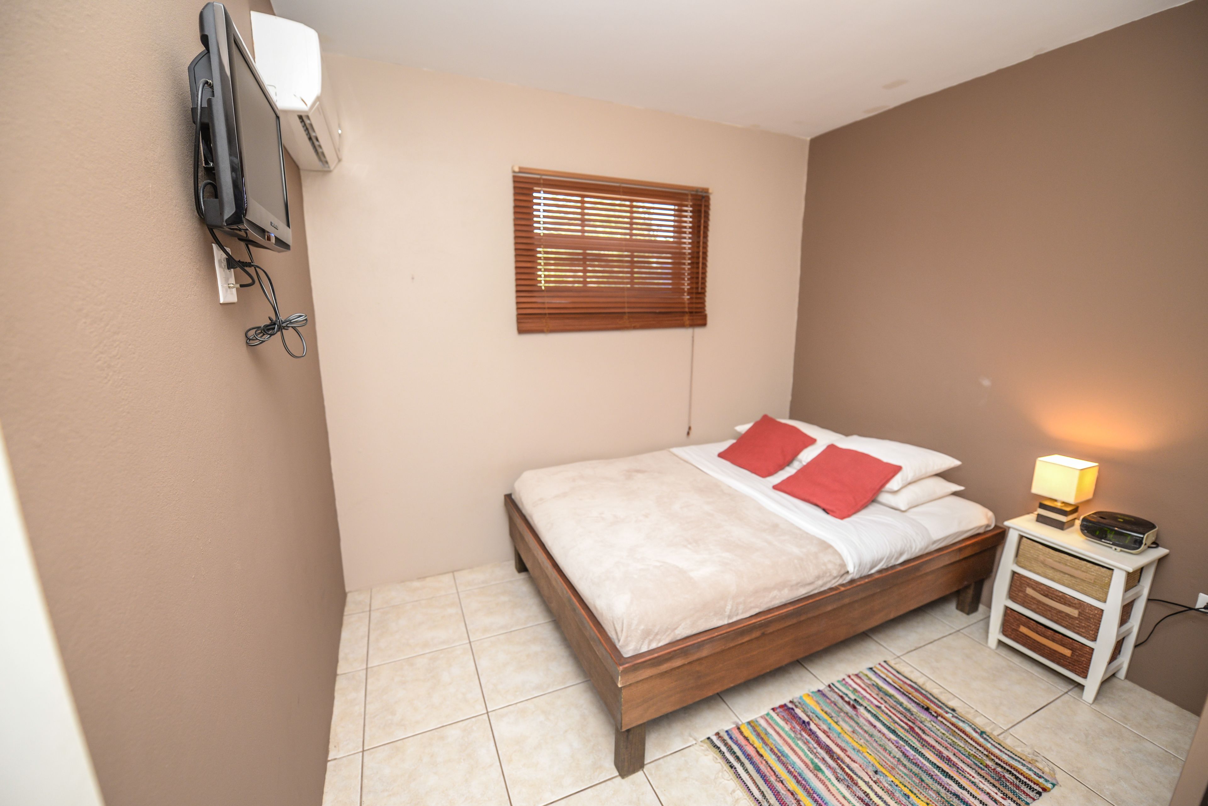 A bedroom with a bed, a bedside table with a lamp, a window with blinds, and an air conditioning unit mounted on the wall.