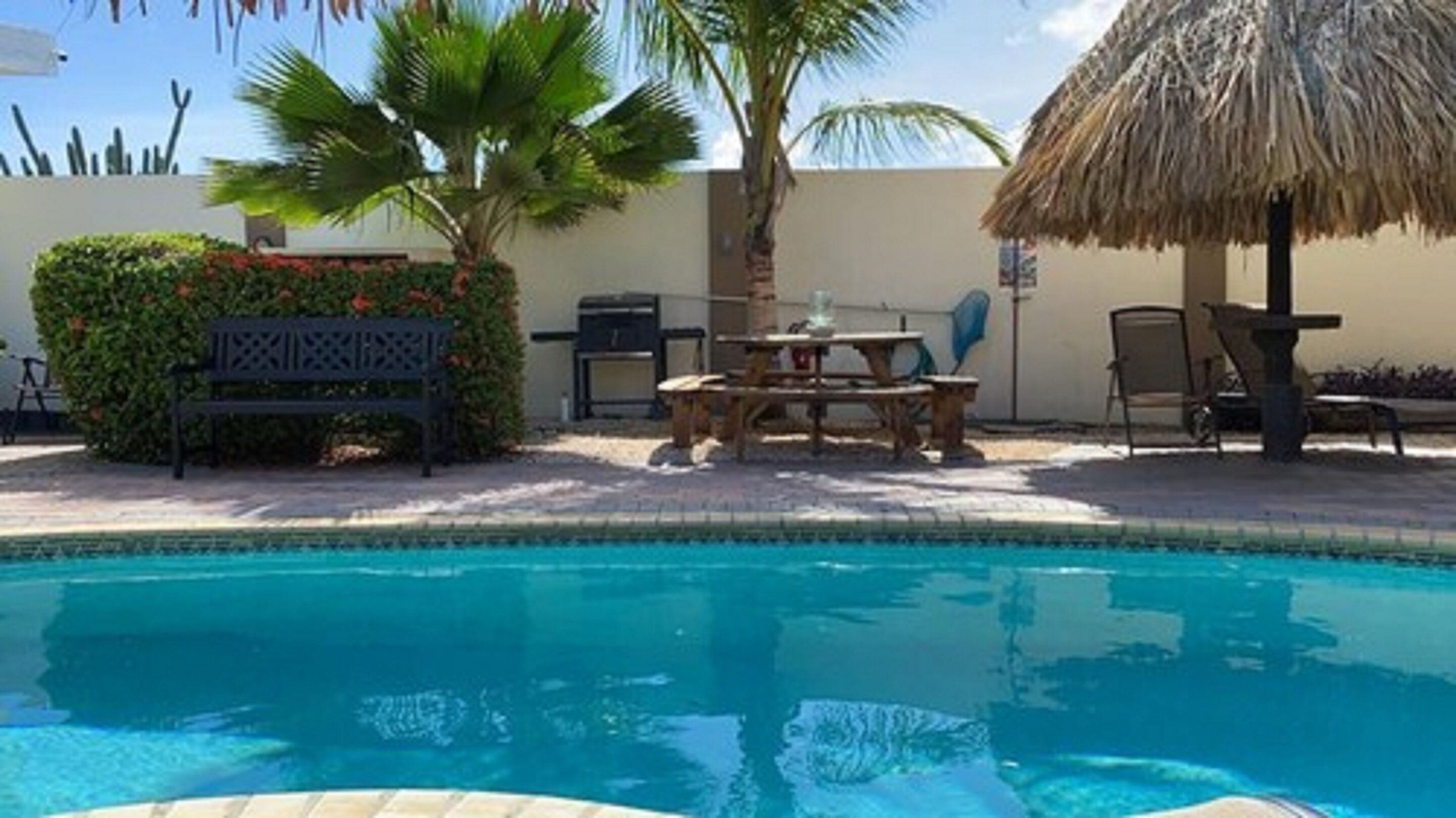 A swimming pool with a thatched umbrella, a bench, and a table with chairs.