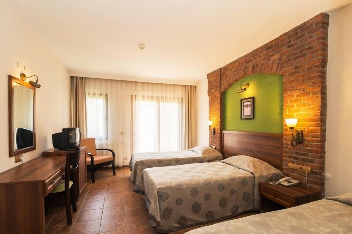 Ett hotellrum med två sängar, ett skrivbord med en tv, en stol, en vägg med synliga tegelstenar och ett fönster med gardiner.