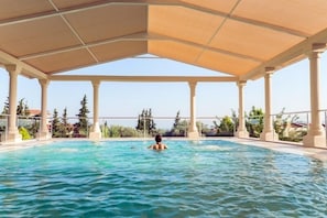 Une piscine couverte à l’eau claire, entourée de colonnes, avec en arrière-plan une vue sur des arbres et des bâtiments.