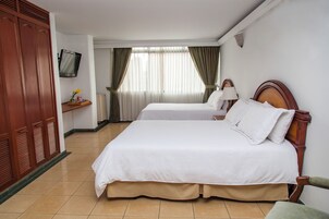 A hotel room with two beds, a TV mounted on the wall, a desk with a lamp, and a wardrobe.