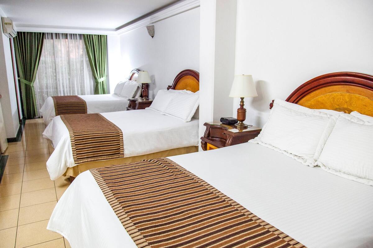 Une chambre d’hôtel avec deux lits, une table de chevet avec une lampe, une commode et une fenêtre avec des rideaux.