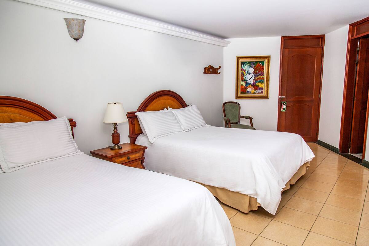 Une chambre d’hôtel avec deux lits simples, une table de chevet avec une lampe, une chaise, une porte en bois et un tableau coloré au mur.