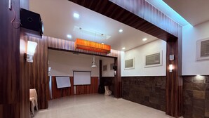 A spacious room with a large open area, a projector screen, and a wooden accent wall. - Hotel Alka Residency (Thane)