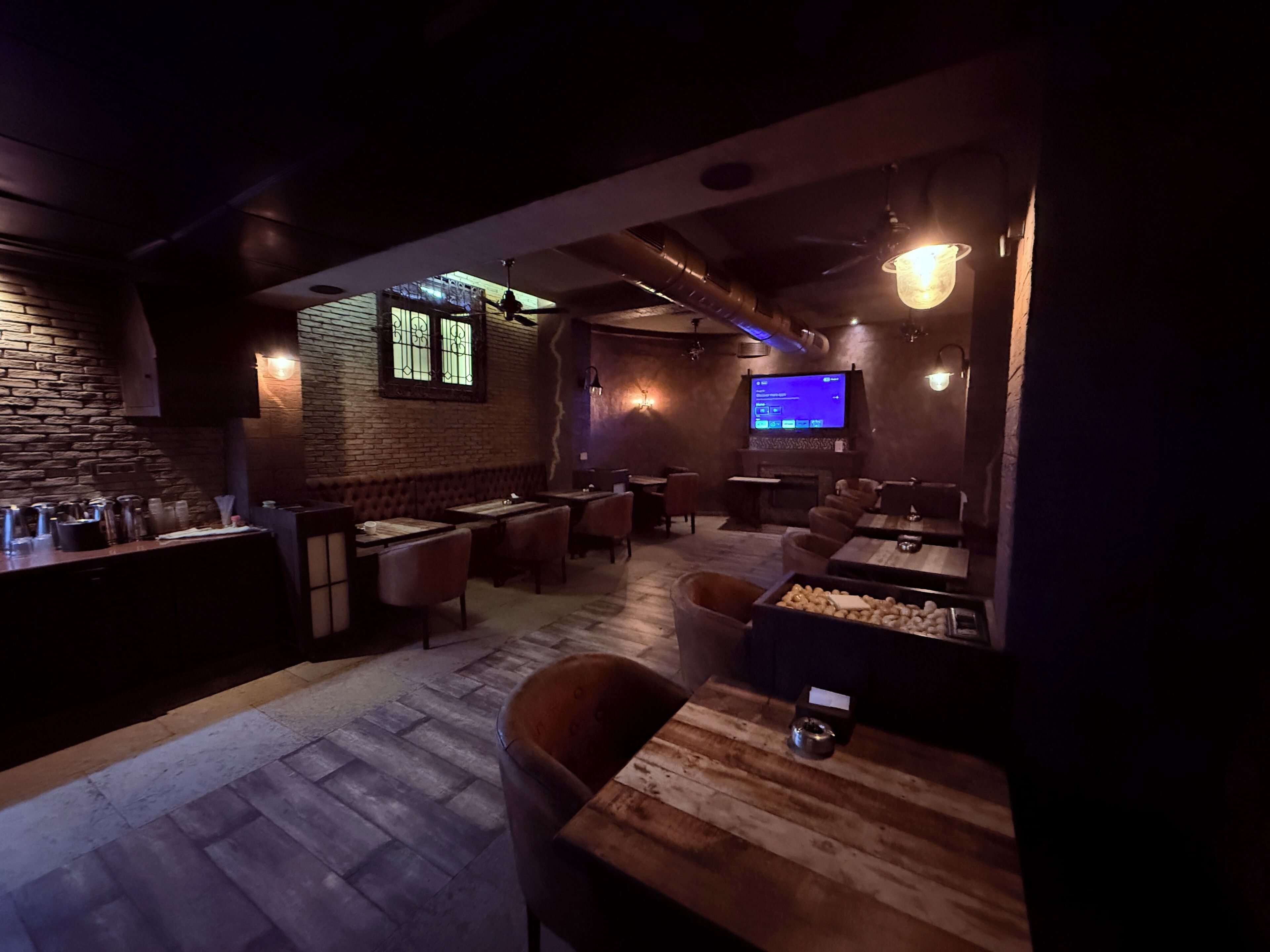 A dimly lit bar area with wooden furniture, a television screen, and a brick wall.