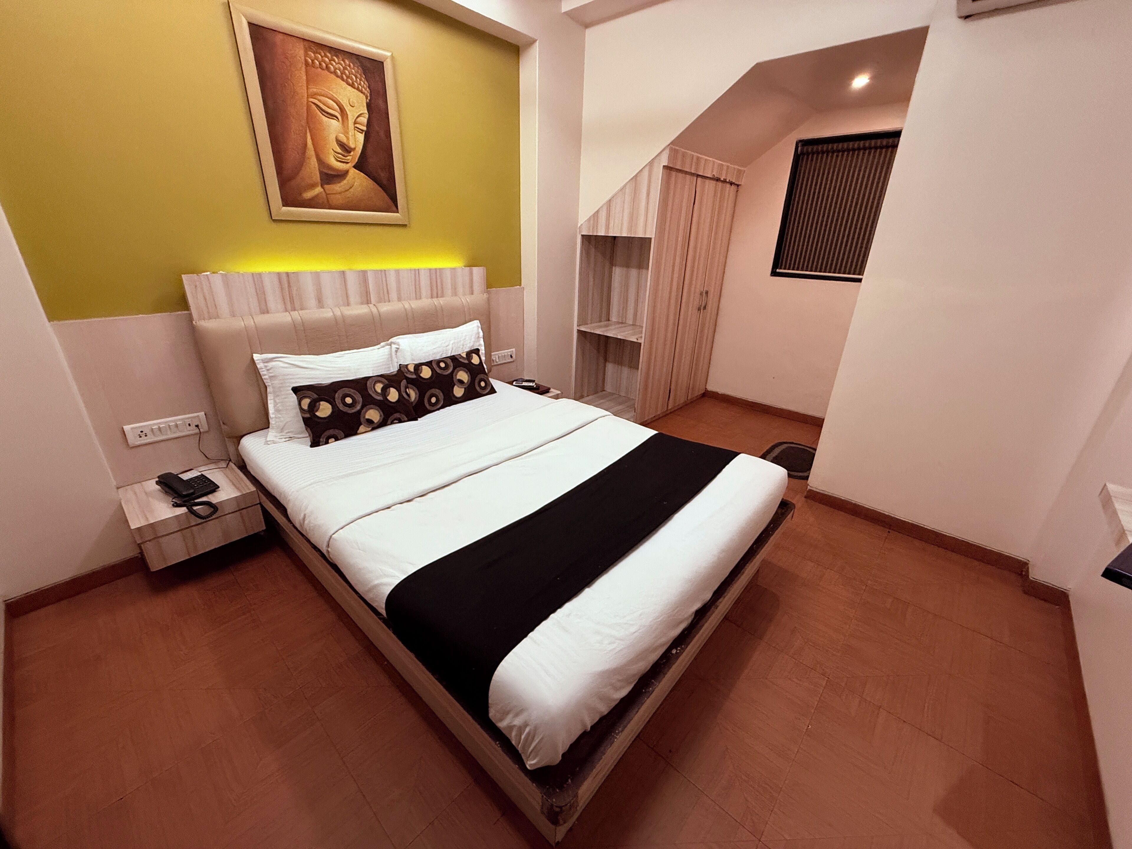 A bedroom with a bed, a nightstand, a wall-mounted Buddha statue, a window with blinds, and a wooden wardrobe.