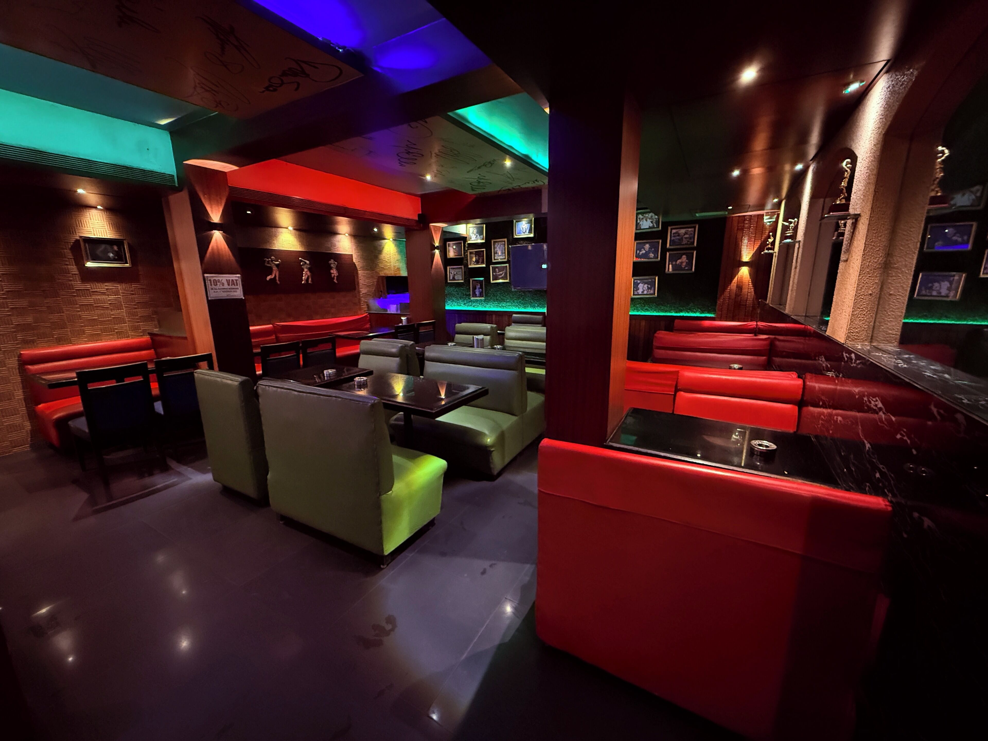 Un espace bar à la lumière tamisée, avec un éclairage d’ambiance rouge et vert, de nombreux types de sièges, notamment des banquettes et des tables, et un mur décoré de tableaux et de trophées encadrés.
