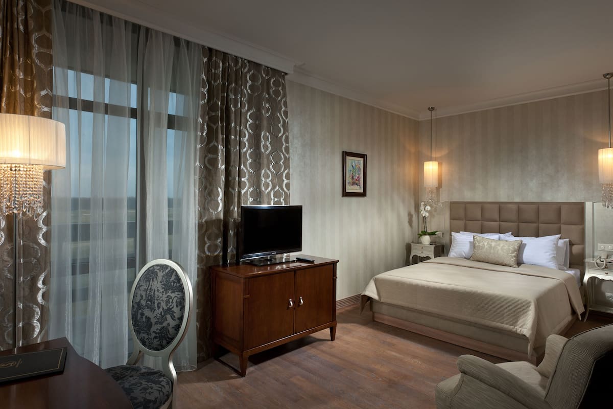 A hotel room with a large bed, a TV on a wooden cabinet, a chair, a desk with a lamp, and curtains.