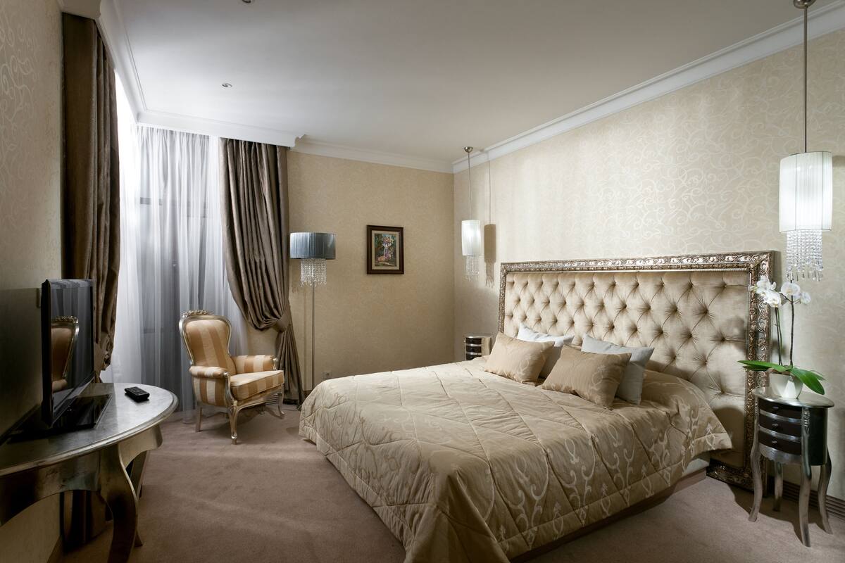 A hotel room with a large bed, a desk with a TV, a chair, and a bedside table with a lamp.
