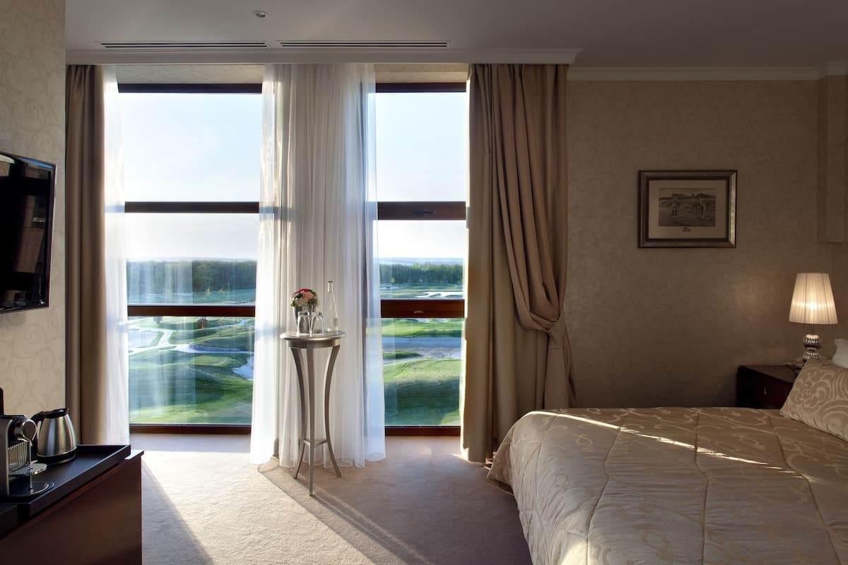 A hotel room with a large window, a bed, a bedside table, a lamp, and a framed picture on the wall.