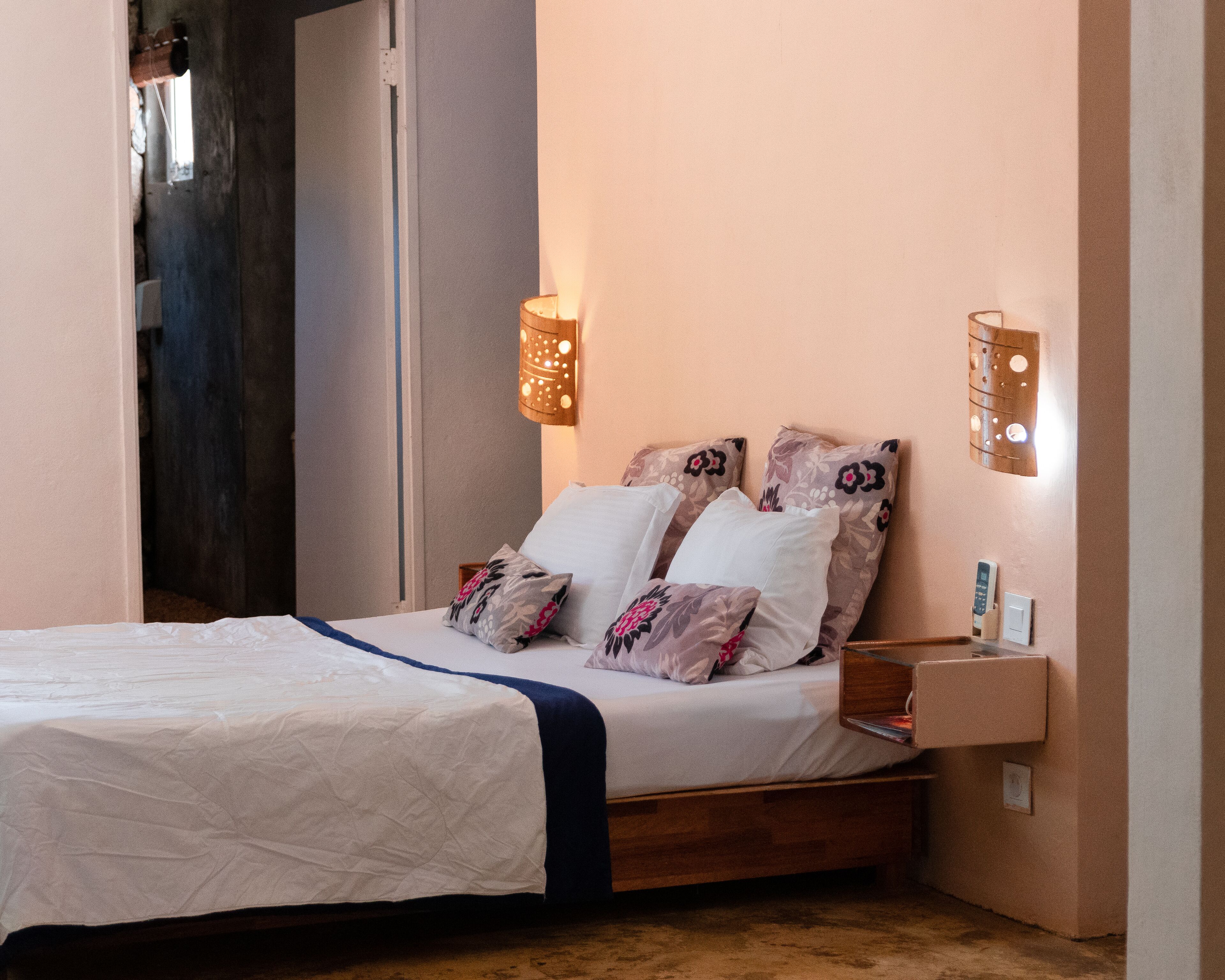 A neatly made bed with a white and blue bedspread, two pillows with floral patterns, and a wooden bedside table with a lamp.