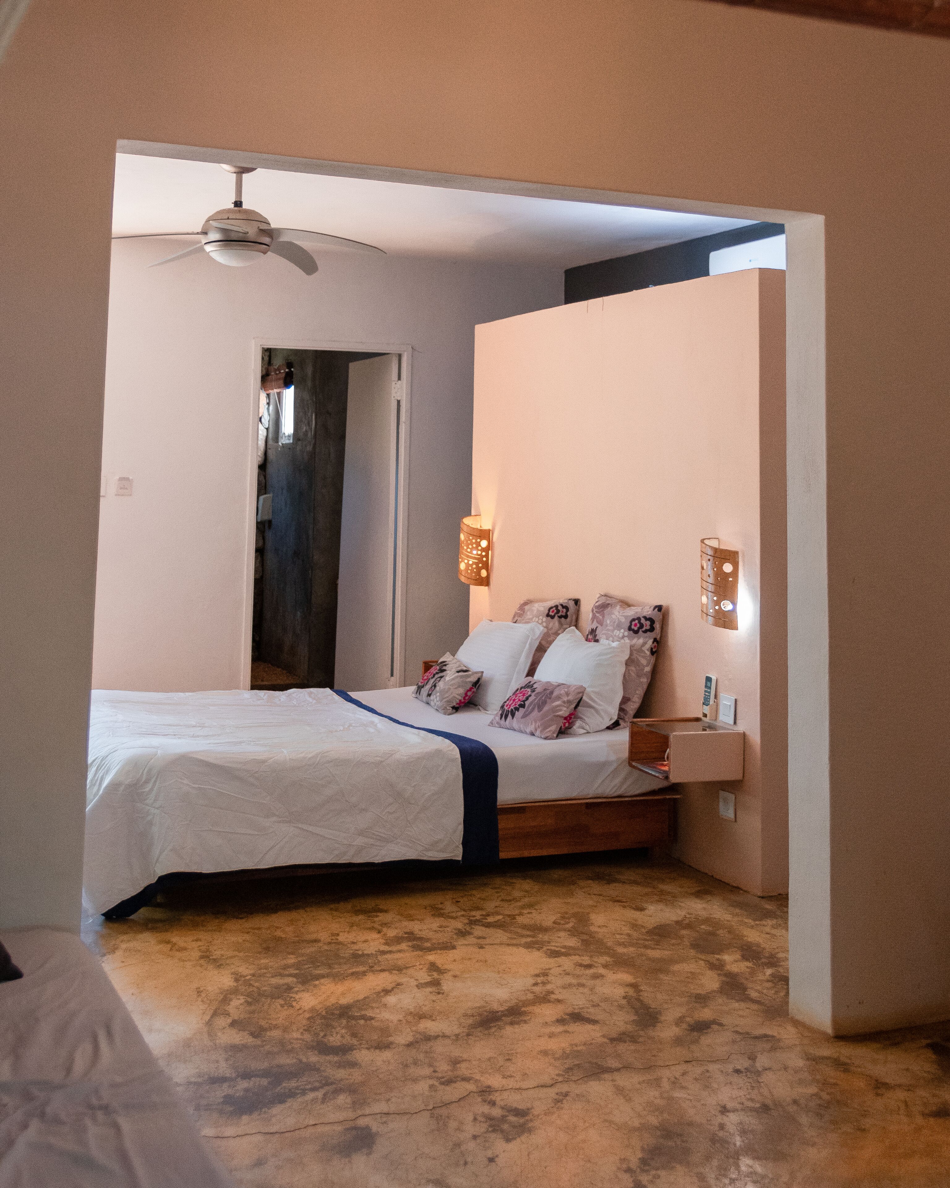 Un dormitorio con una cama, un ventilador de techo, una mesita de noche con lámpara y una puerta que da a otra habitación.