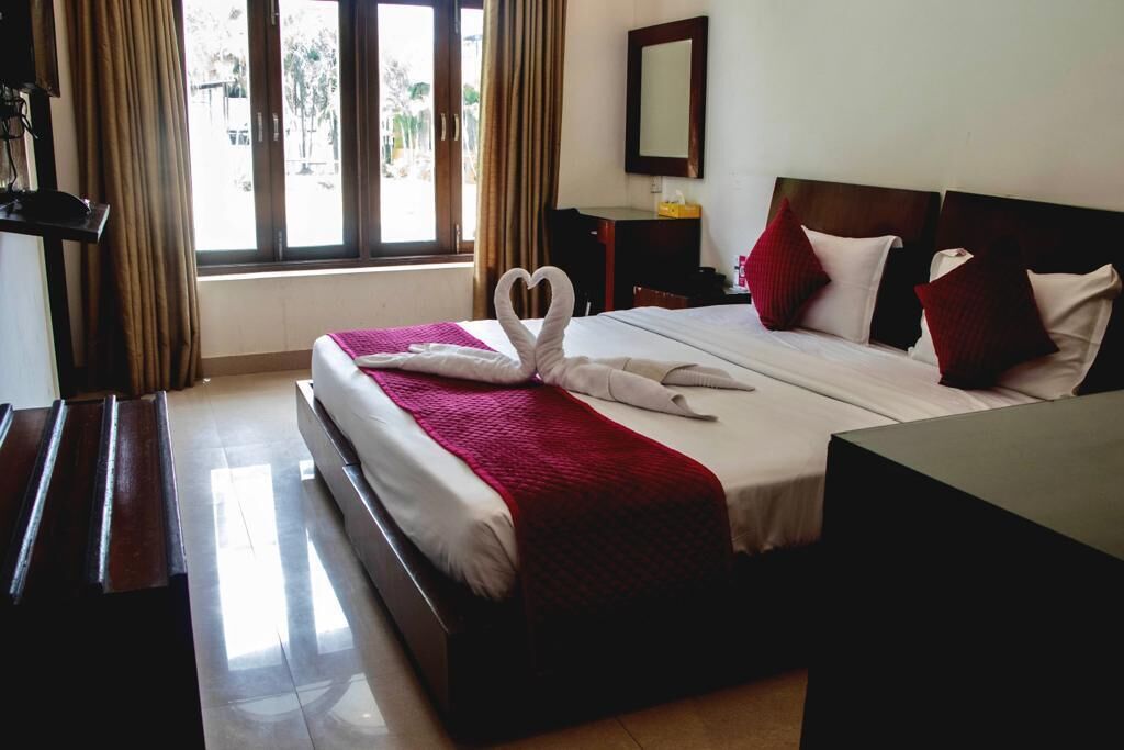 A hotel room with a large bed, a wooden nightstand, a desk, and a window with curtains.