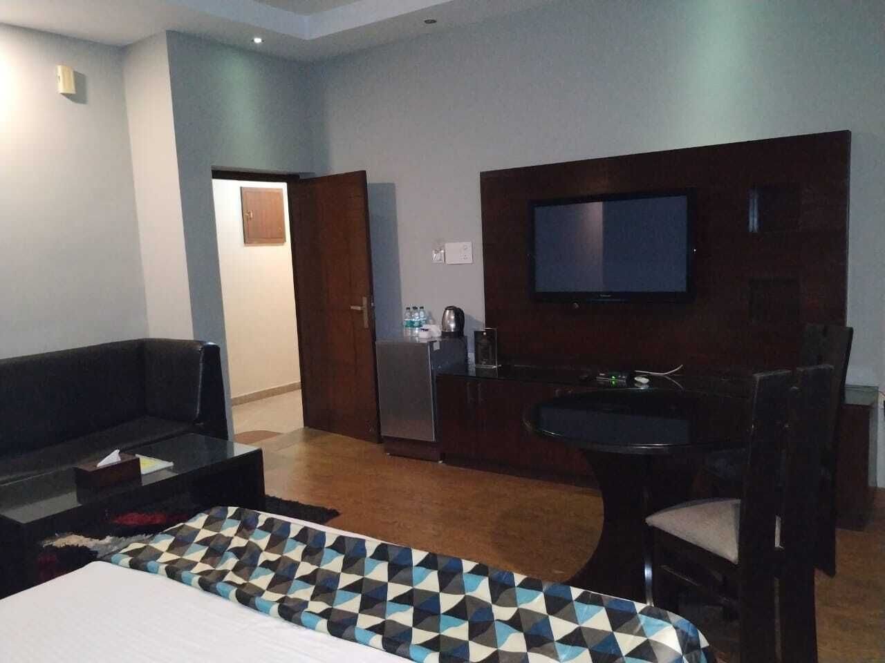 A hotel room with a bed, a sofa, a dining table with chairs, a TV mounted on the wall, and a refrigerator.