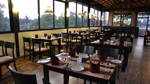 Un restaurante con varias mesas y sillas, amplias ventanas con vistas a zonas verdes e iluminación colgante.