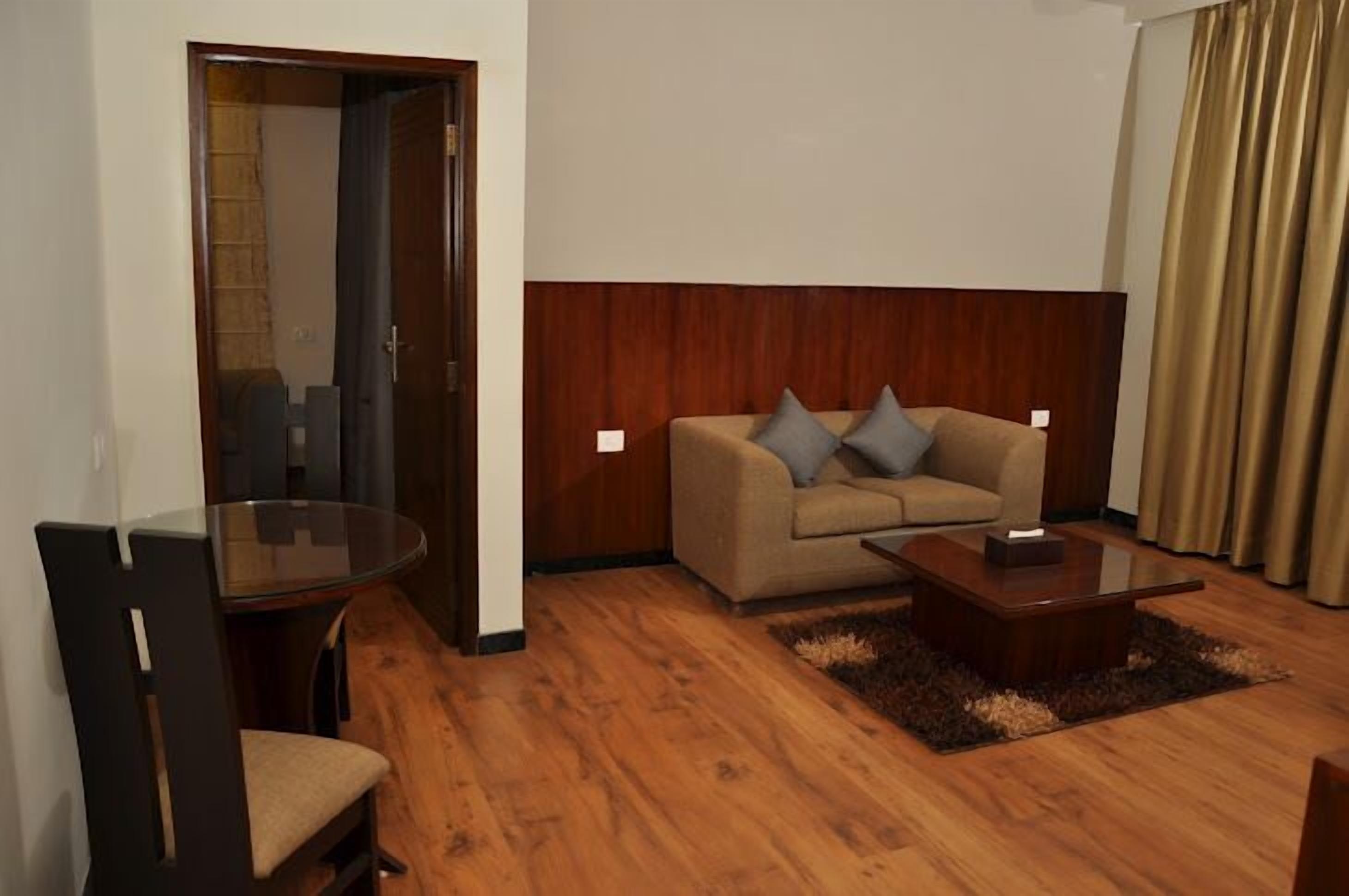 Een kamer met een houten vloer, een beige bank met twee kussens, een glazen tafel en een donkere houten stoel.