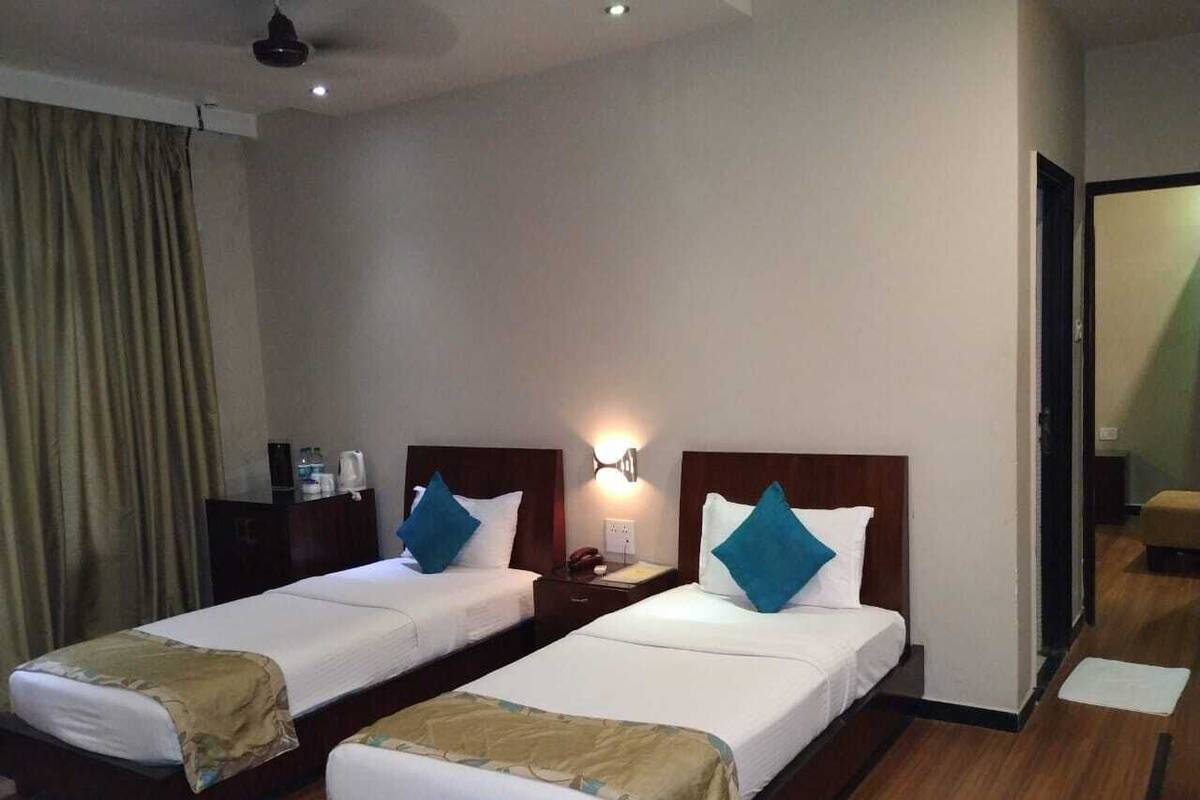 A hotel room with two single beds, a nightstand with a lamp, a wooden floor, and a view of the hallway.