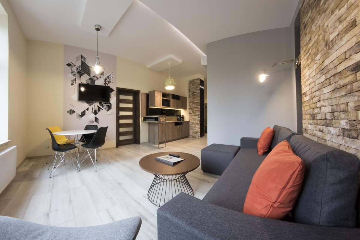 Una sala de estar moderna con un sofá gris, cojines anaranjados, una mesa de centro redonda y un comedor con una mesa blanca y sillas negras.