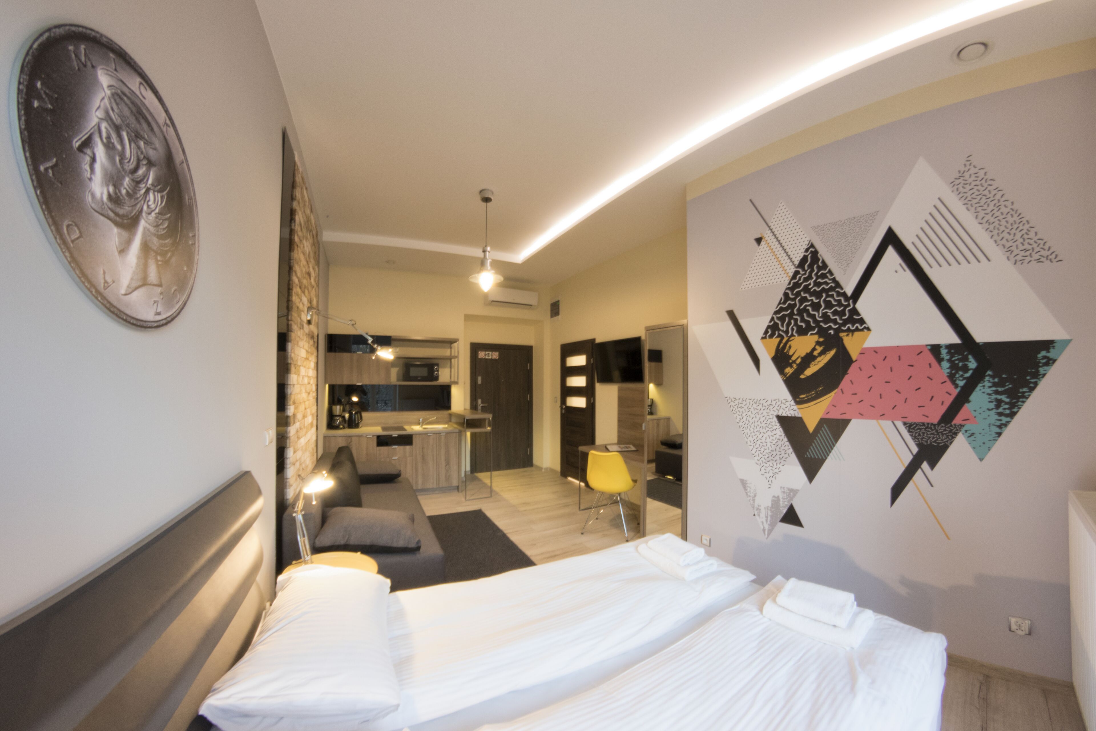 A modern hotel room with a neatly made bed, a dining area, and a kitchen in the background.