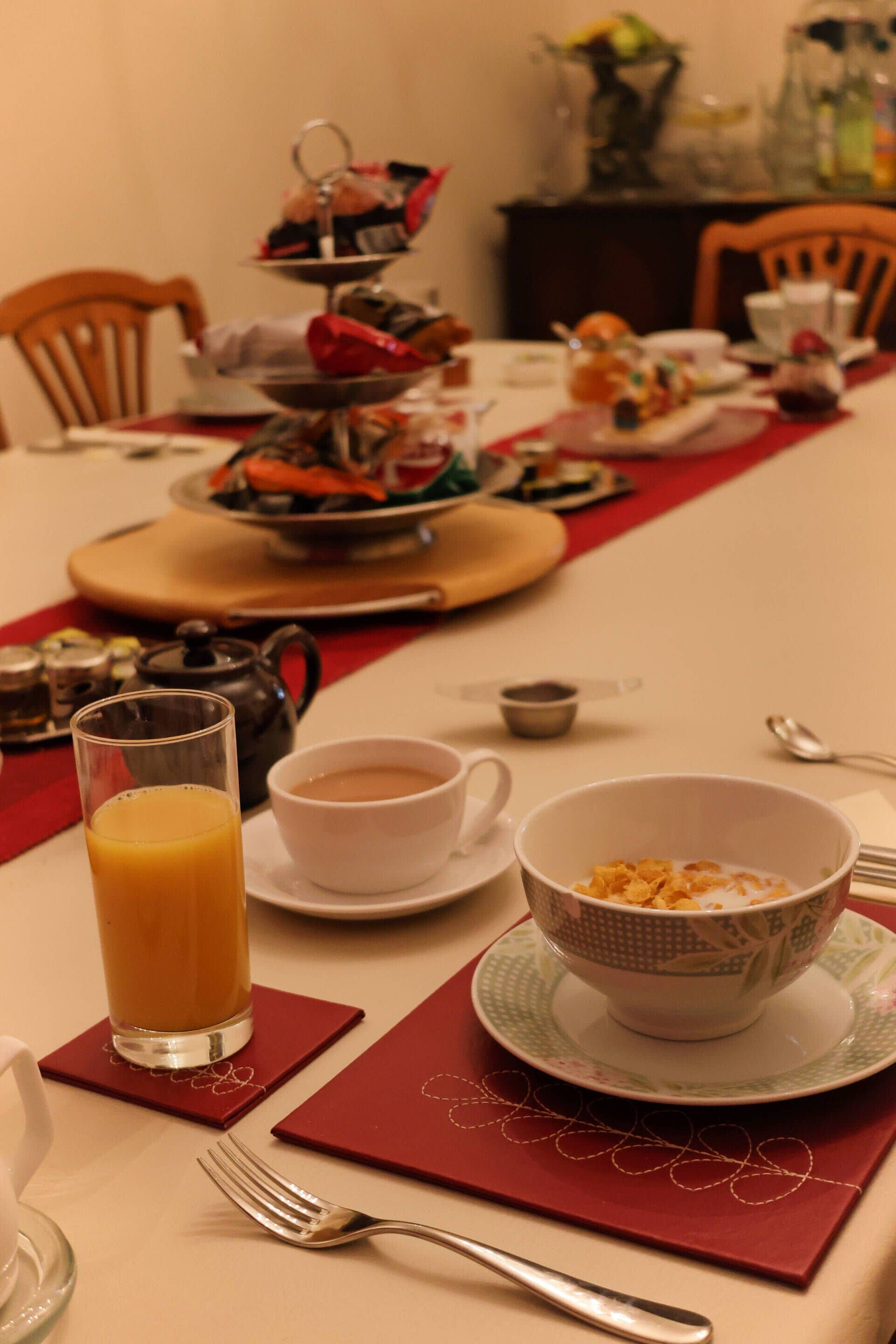 A dining table set with breakfast items including a glass of orange juice, a cup of tea, a bowl of cereal, and a teapot.