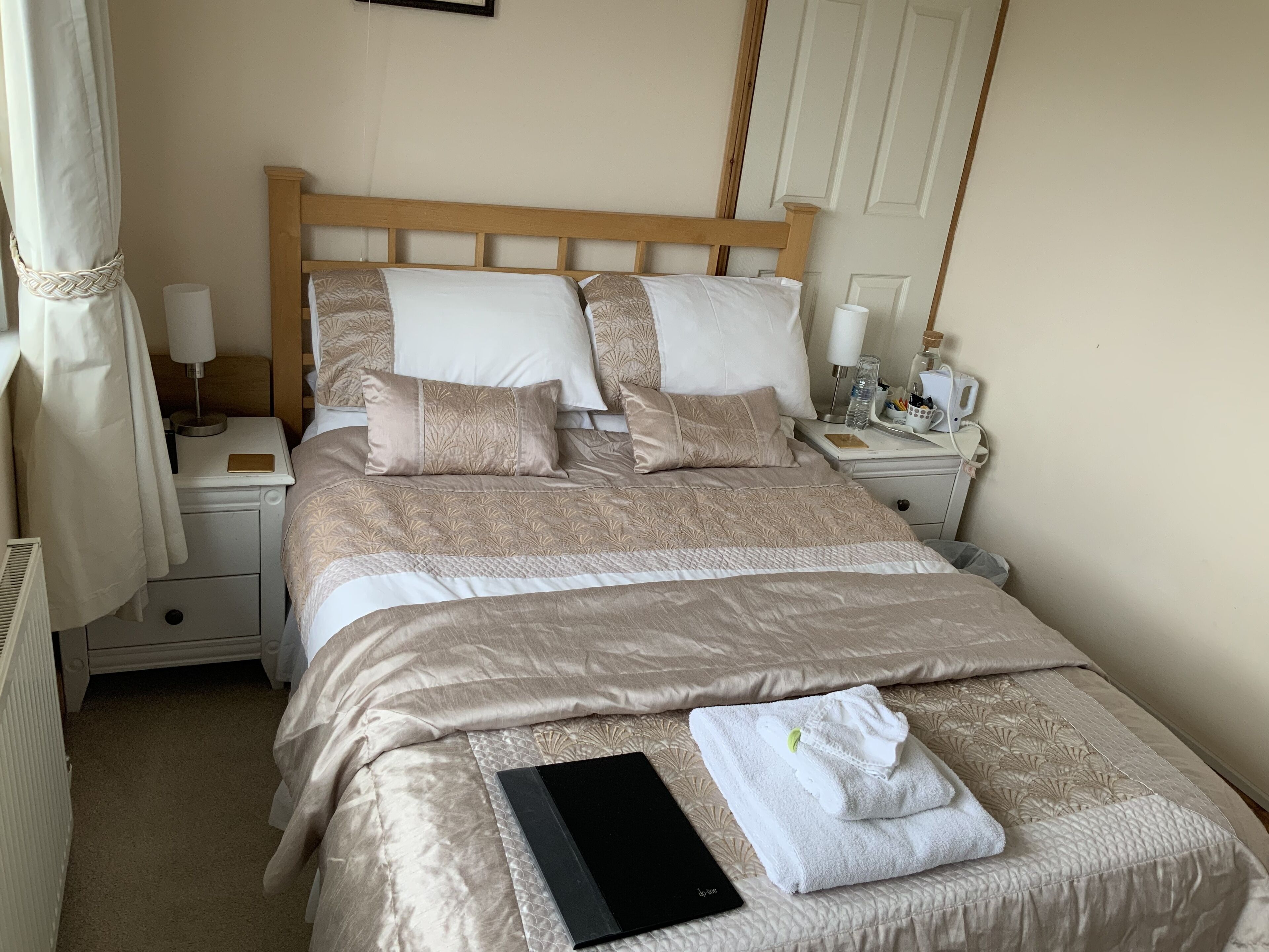 A neatly made bed with a patterned bedspread, pillows, and a folded towel on the bed. There is a bedside table with a lamp and a bottle of water.