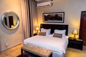 Une chambre d’hôtel avec un grand lit, deux tables de chevet avec des lampes, un miroir rond, un tableau encadré au mur et un appareil de climatisation.