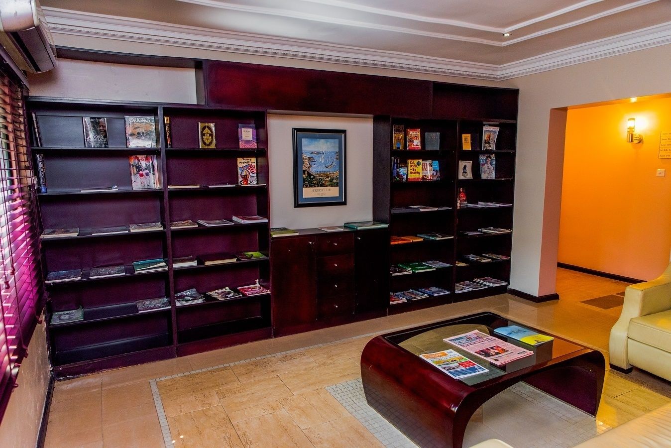 Une bibliothèque avec des étagères, une table basse avec des magazines et un tableau encadré au mur.