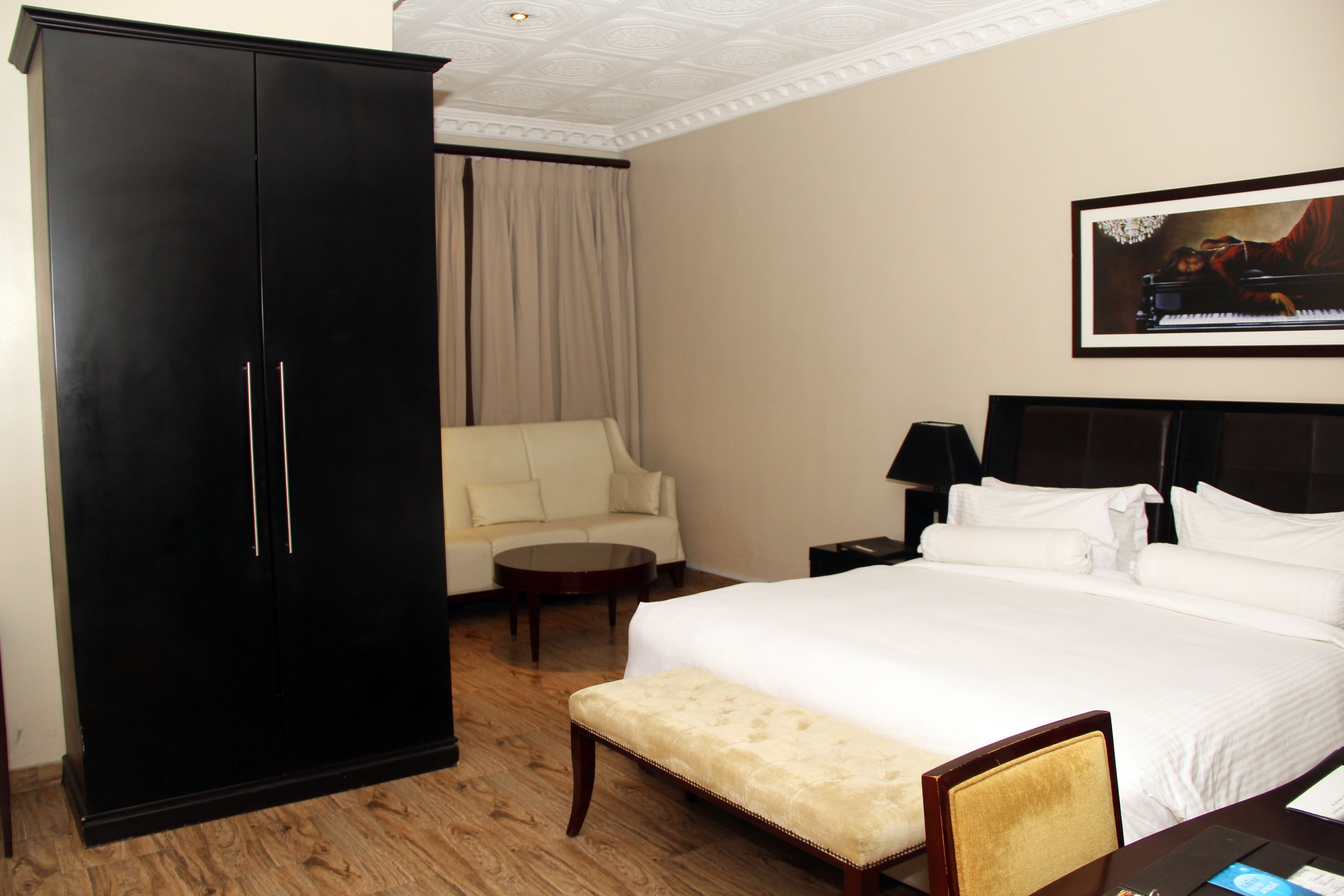 A hotel room with a large bed, a sitting area with a chair and a small table, a wardrobe, and a painting on the wall.