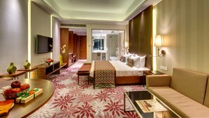 A modern hotel room with a large bed, a sofa, a flat-screen TV, a desk, and a dining table with fruit and a gift box. - Pride Plaza Hotel, Kolkata (Barasat)