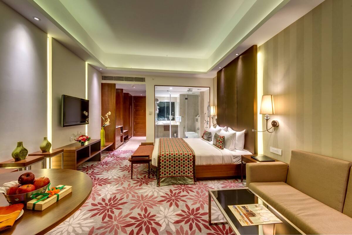 A modern hotel room with a large bed, a sofa, a flat-screen TV, a desk, and a dining table with fruit and a gift box.