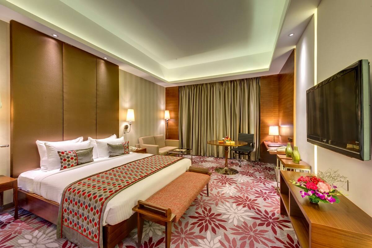 A hotel room with a large bed, a bench, a desk, a chair, a TV, and a table with lamps.