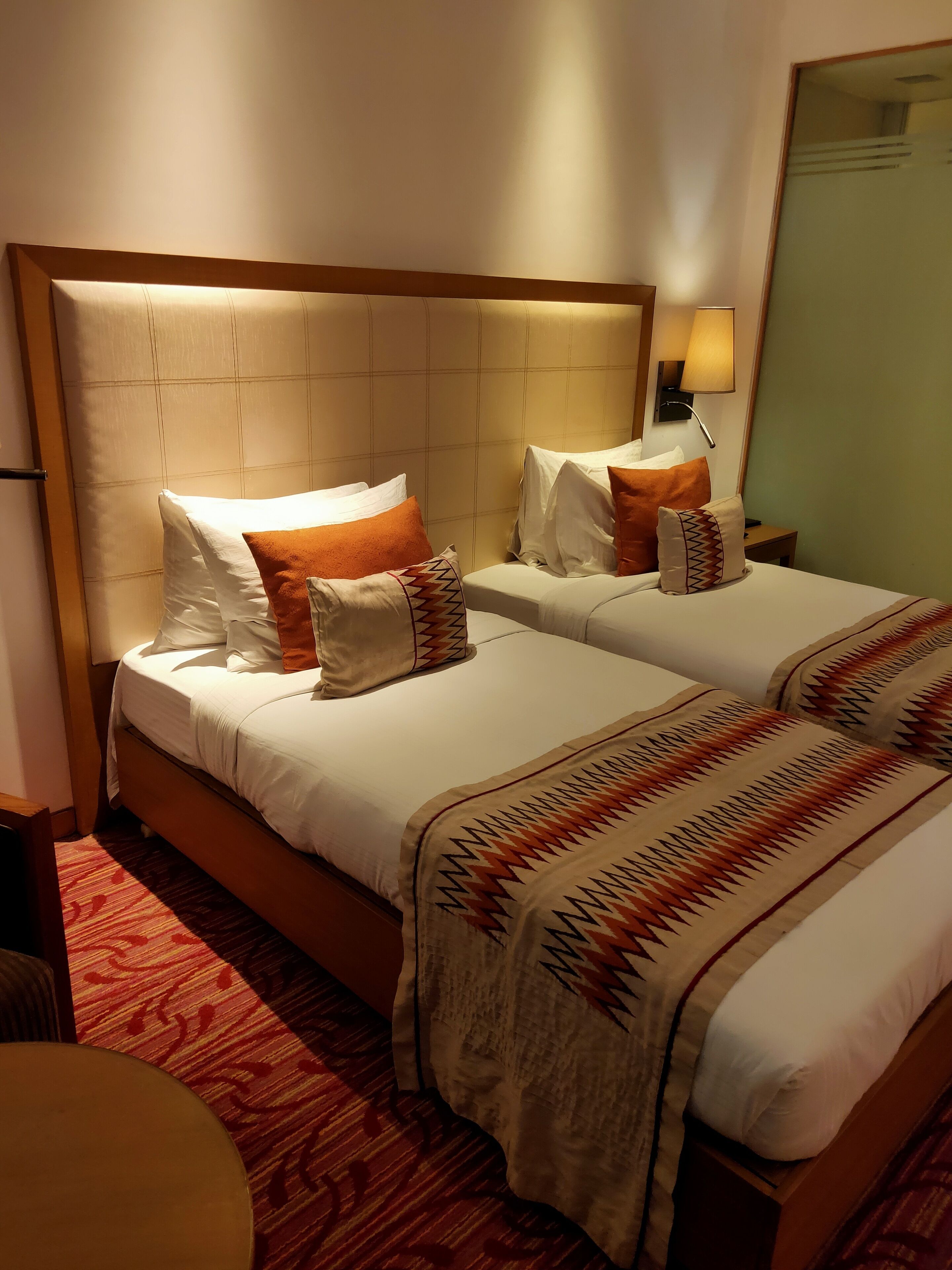 Habitación de hotel con dos camas, cada una con ropa blanca y colchas decorativas. Se observa un cabecero de madera, una mesita de noche con lámpara, y un espejo grande en la pared.