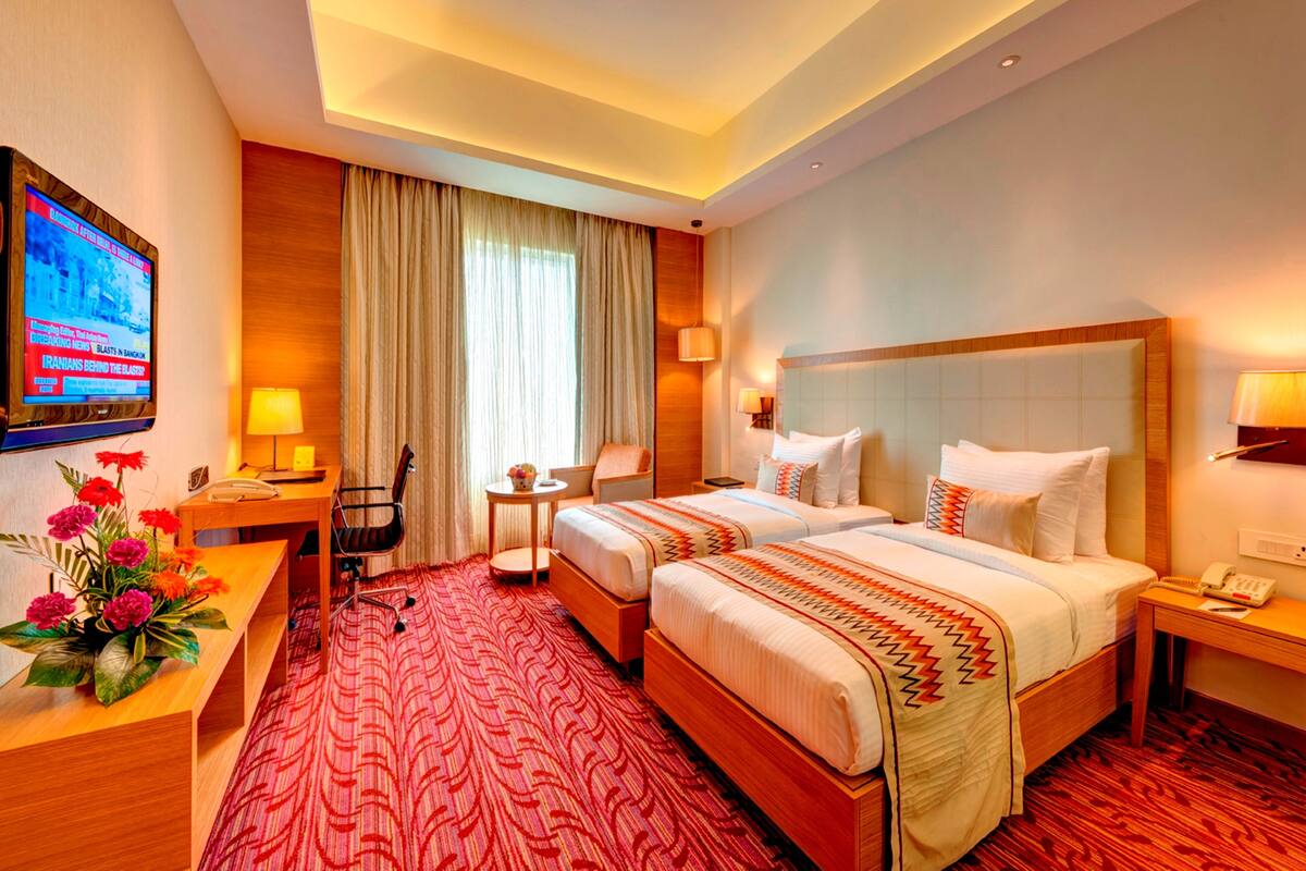 A hotel room with two beds, a desk, a chair, a TV, and a red patterned carpet.