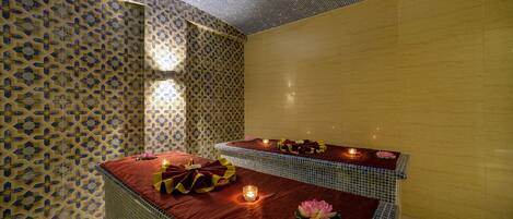 A spa room with patterned walls, two massage tables with red linens, candles, and lotus flowers.
