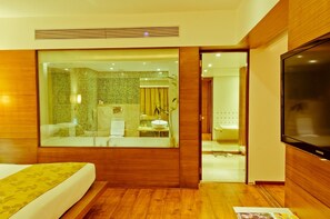 A modern hotel room with a large bed, a TV, a bathroom with a shower, and a view through a glass partition. - Ramee Grand Hotel and Spa, Pune (Pune)