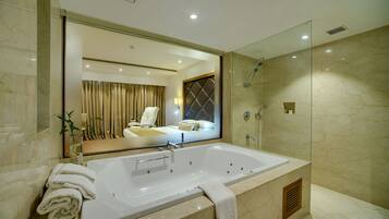 A modern bathroom with a large bathtub, a glass shower enclosure, and a mirror reflecting a bedroom.