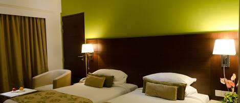 A hotel room with two beds, a desk, a chair, and a nightstand with a lamp and flowers.