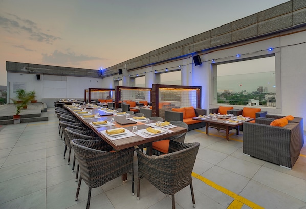 A rooftop dining area with long tables, wicker chairs, and orange cushions.