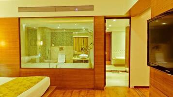 Habitación de hotel con una cama grande, televisión, baño con ducha y lavamanos, y vistas a través de un panel de vidrio.